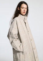 A person with long, wavy brown hair poses against a light grey background, wearing the TYPE-O 002 coat by A-POC ABLE ISSEY MIYAKE—a light beige grid-patterned piece with a high collar made from lightweight stretch material.