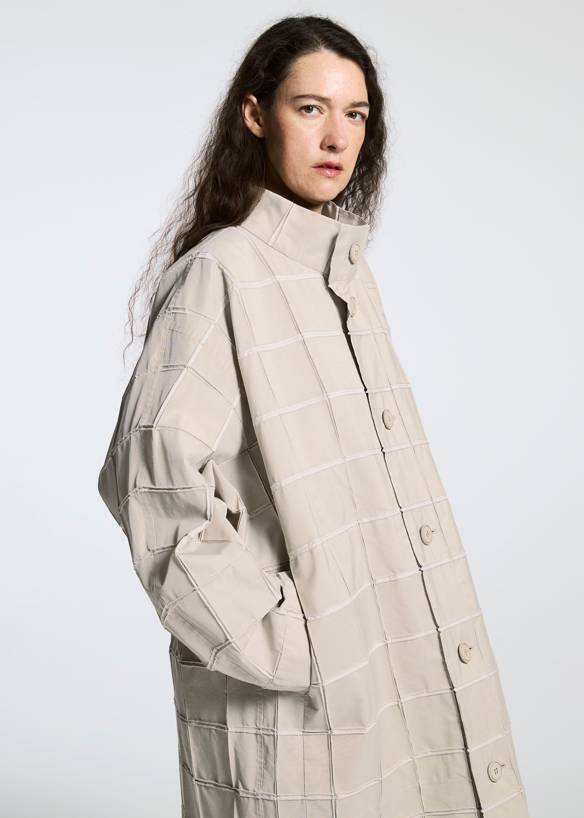 Zoomed view A person with long, wavy brown hair poses against a light grey background, wearing the TYPE-O 002 coat by A-POC ABLE ISSEY MIYAKE—a light beige grid-patterned piece with a high collar made from lightweight stretch material.