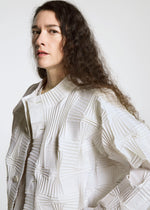 A person with long, wavy brown hair models the TYPE-O 018 pleated jacket by A-POC ABLE ISSEY MIYAKE, featuring geometric patterns, while standing against a plain light background and gazing slightly to the side.