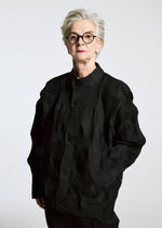 An older person with short gray hair and round glasses stands confidently in the TYPE-O 018 black pleated jacket by A-POC ABLE ISSEY MIYAKE, paired with dark pants against a plain light background.