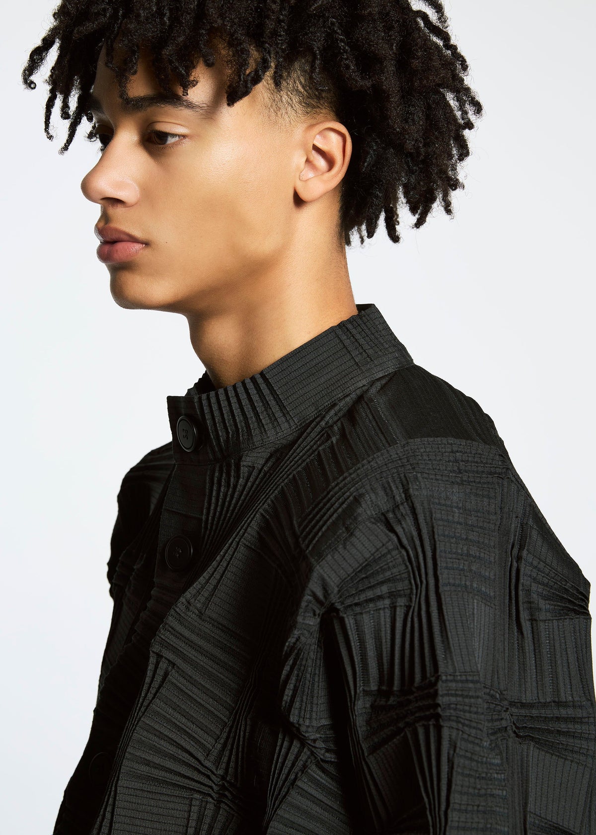 A person with short, curly hair wears the TYPE-O 018 pleated black jacket by A-POC ABLE ISSEY MIYAKE, featuring a high collar and large buttons, looking left against a plain white background. view 8 of 8