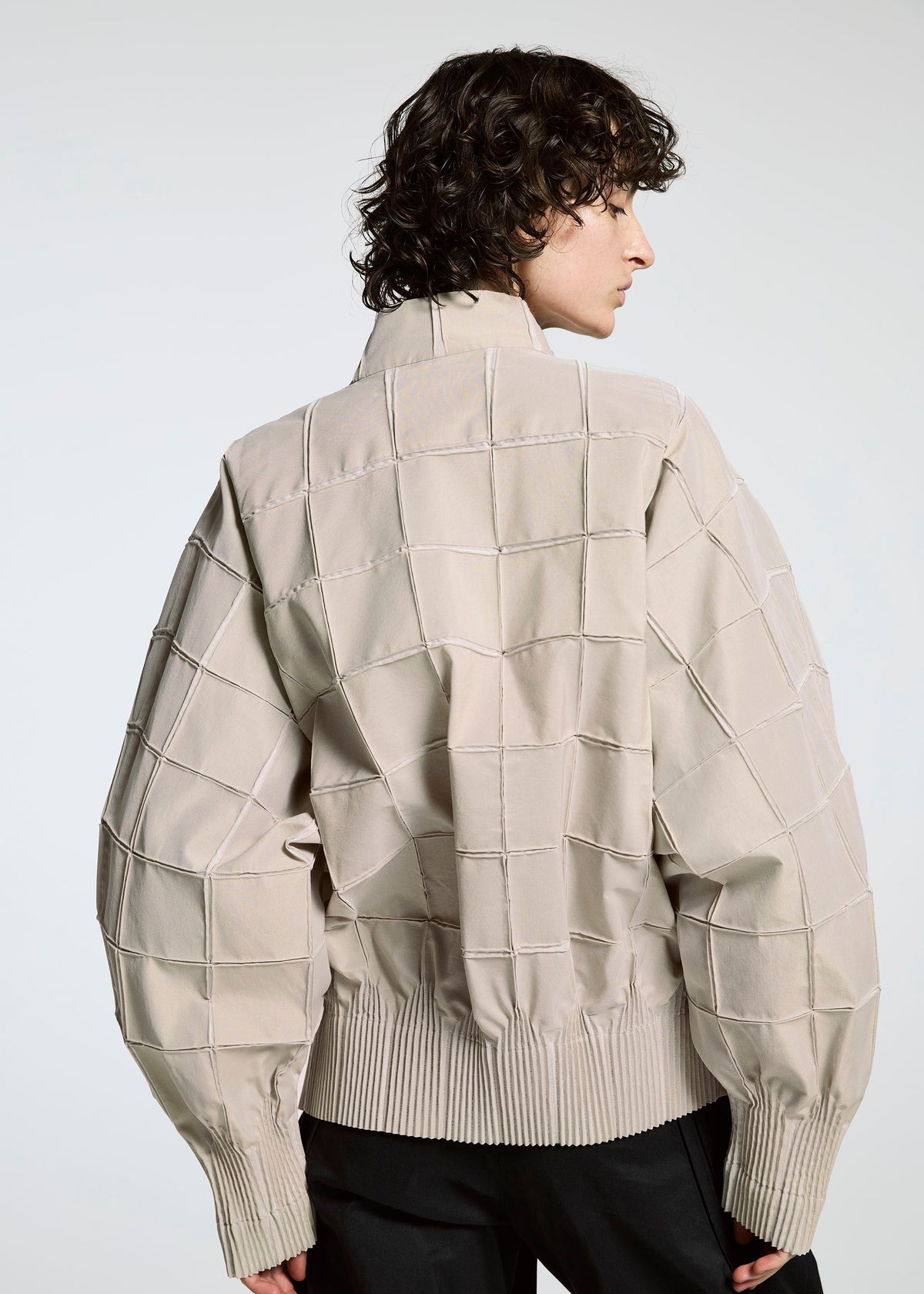 A person with curly hair is seen from behind, wearing the TYPE-O 002 jacket by A-POC ABLE ISSEY MIYAKE—a light beige grid-patterned piece with wide sleeves and ribbed cuffs and hem, made of lightweight stretch material. The background is plain and light-colored. view 6 of 9