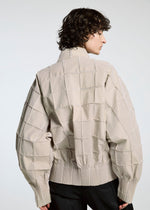 A person with curly hair is seen from behind, wearing the TYPE-O 002 jacket by A-POC ABLE ISSEY MIYAKE—a light beige grid-patterned piece with wide sleeves and ribbed cuffs and hem, made of lightweight stretch material. The background is plain and light-colored.