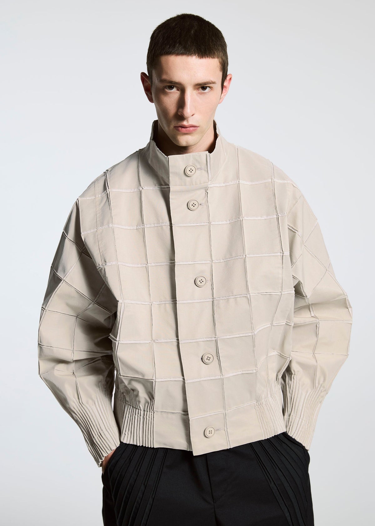 A person with short brown hair wears the TYPE-O 002 jacket by A-POC ABLE ISSEY MIYAKE, featuring a grid pattern, large buttons, ribbed cuffs, and lightweight stretch fabric, standing against a plain light background. view 7 of 9