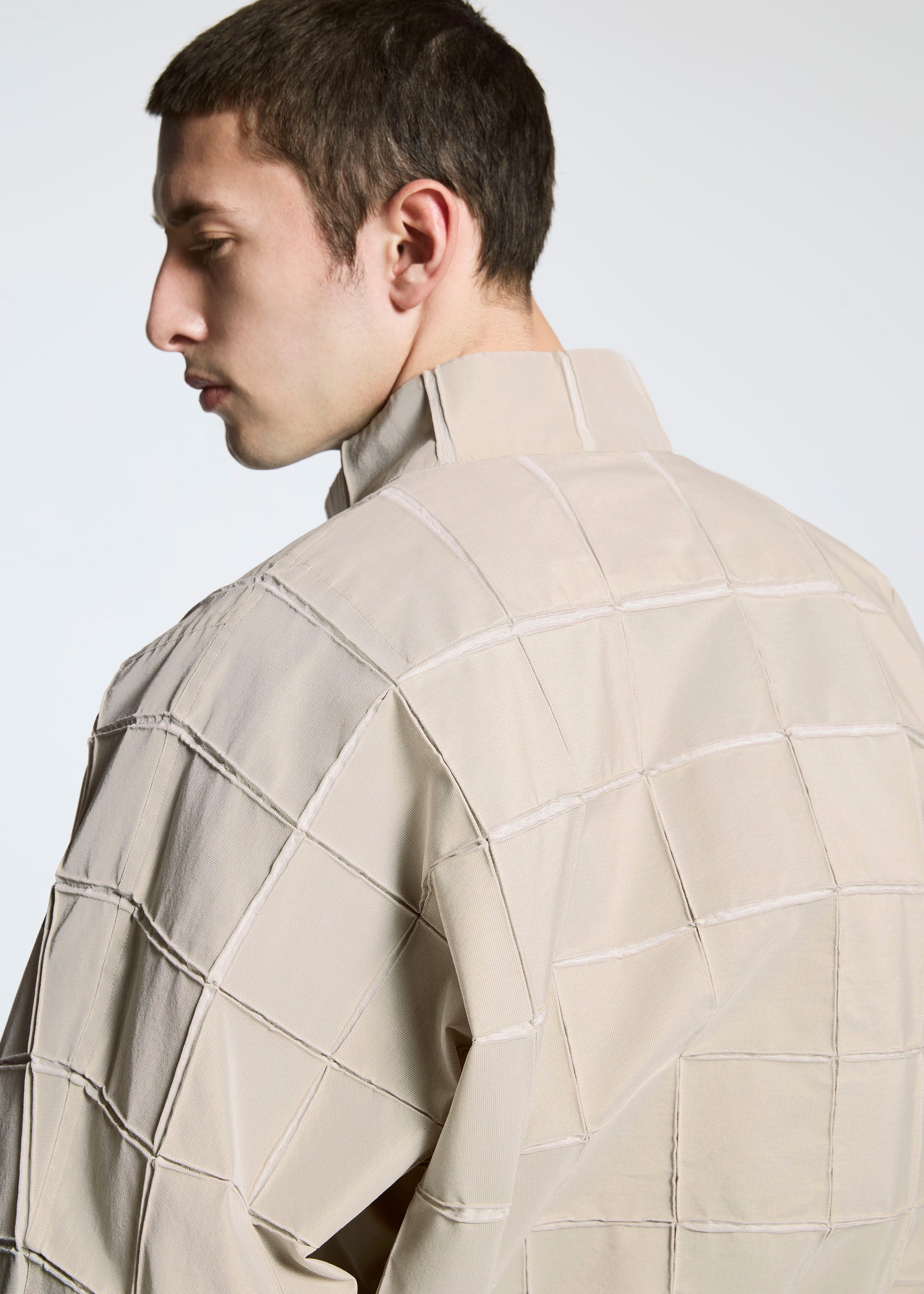 Zoomed view A young man with short dark hair is shown from the side and back, wearing the TYPE-O 002 grid pattern jacket by A-POC ABLE ISSEY MIYAKE, made from lightweight stretch material, set against a plain light background.