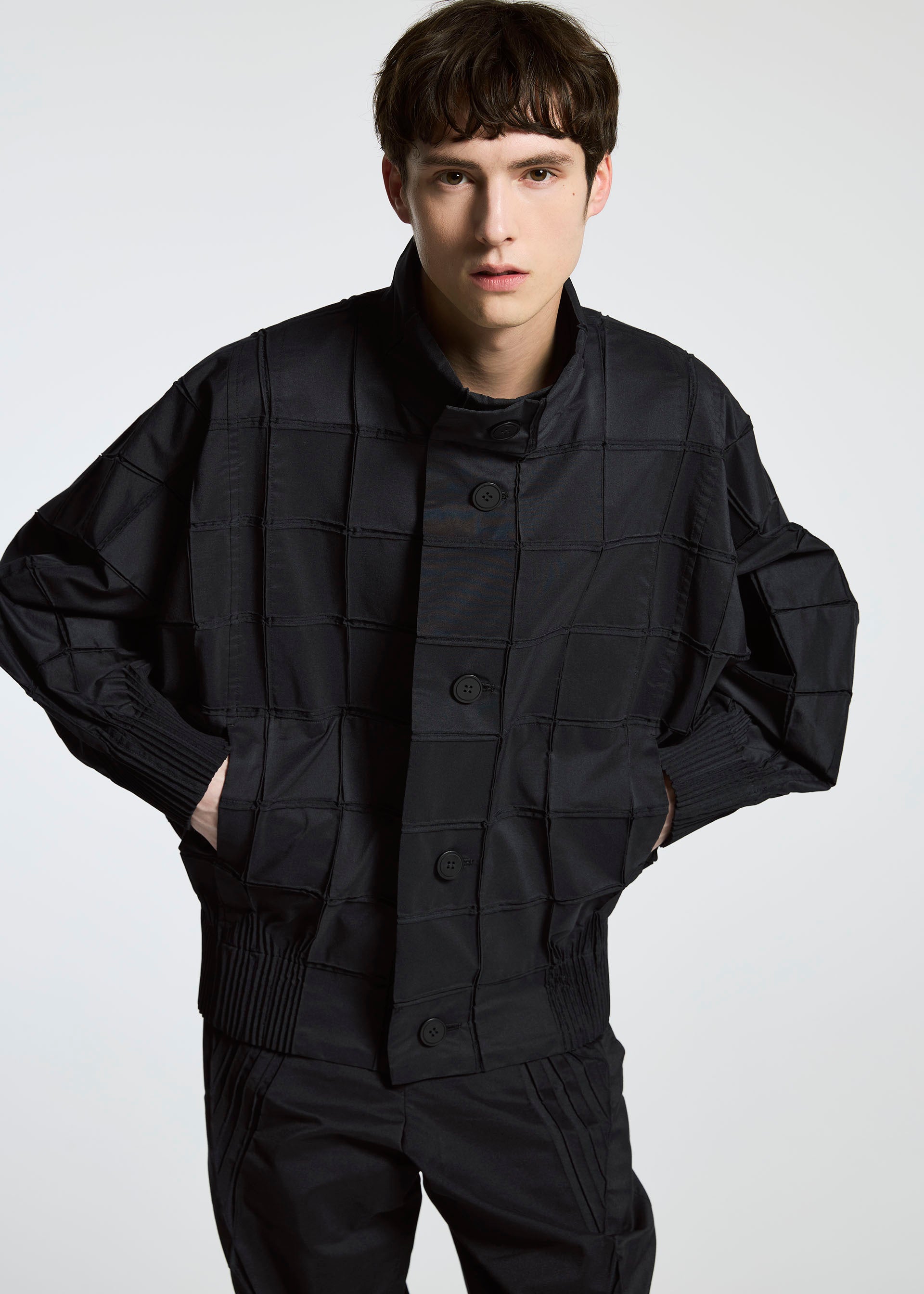 Zoomed view A young person with short brown hair wears the TYPE-O 002 jacket and matching pants by A-POC ABLE ISSEY MIYAKE, featuring a loose fit and grid pattern in lightweight stretch material, standing against a plain light background with hands in pockets.