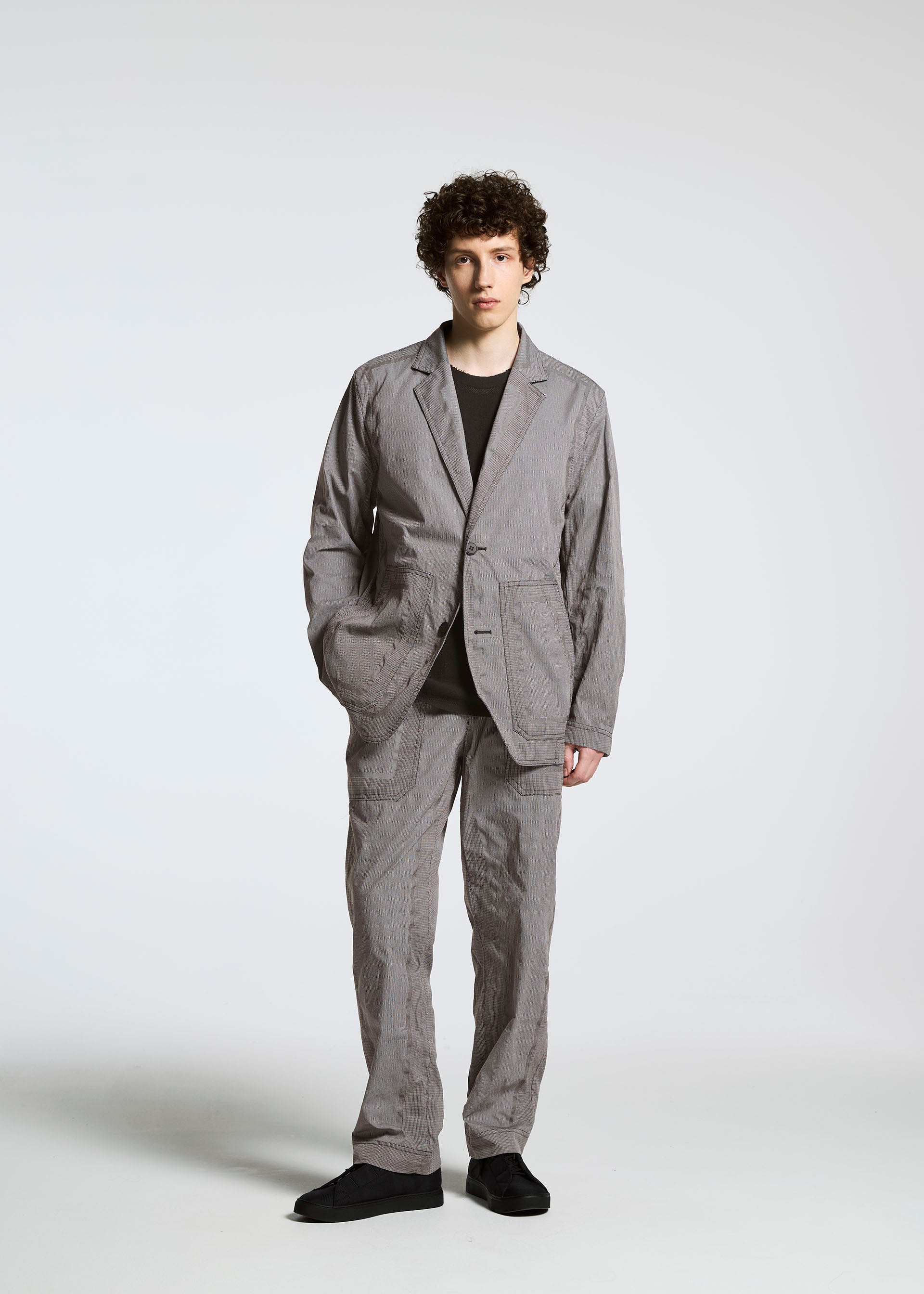 Zoomed view A person with curly hair stands against a plain background wearing the TYPE-U 001 jacket by A-POC ABLE ISSEY MIYAKE, paired with a black shirt and shoes, hands in pockets, looking at the camera.