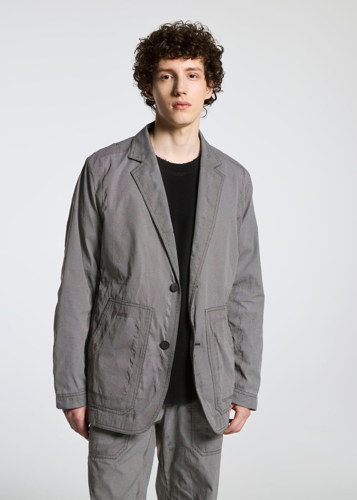A person with curly brown hair wears the TYPE-U 001 relaxed-fit gray blazer and matching pants by A-POC ABLE ISSEY MIYAKE over a black shirt, standing against a plain white background. view 5 of 9