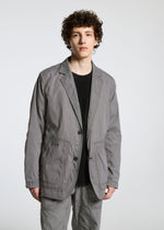 A person with curly brown hair wears the TYPE-U 001 relaxed-fit gray blazer and matching pants by A-POC ABLE ISSEY MIYAKE over a black shirt, standing against a plain white background.