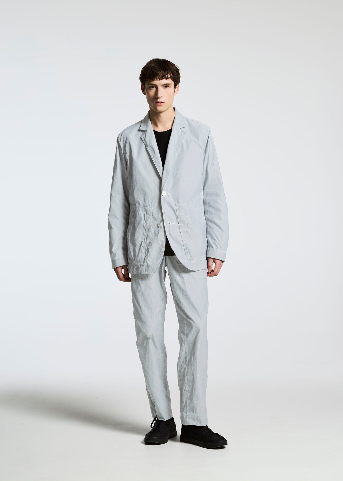 A person with short, dark hair and a neutral expression stands facing forward against a plain white background, wearing the TYPE-U 001 jacket by A-POC ABLE ISSEY MIYAKE, styled with a black shirt and black shoes. view 6 of 9