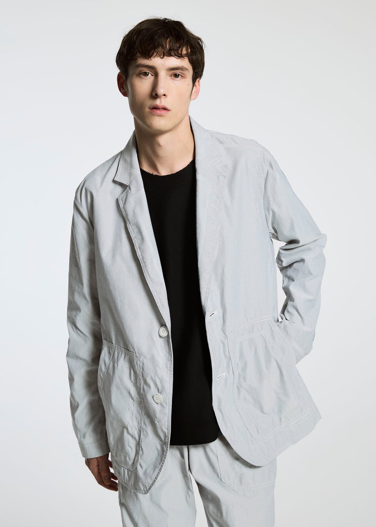 A young man with short brown hair models the TYPE-U 001 relaxed fit light gray jacket and matching pants by A-POC ABLE ISSEY MIYAKE, layered over a black shirt against a plain white background. view 7 of 9