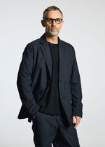 A man with short hair and a beard models the TYPE-U 001 from A-POC ABLE ISSEY MIYAKE—black glasses, wrinkle-resistant jacket, black shirt, and matching pants—standing before a plain light background, looking at the camera.