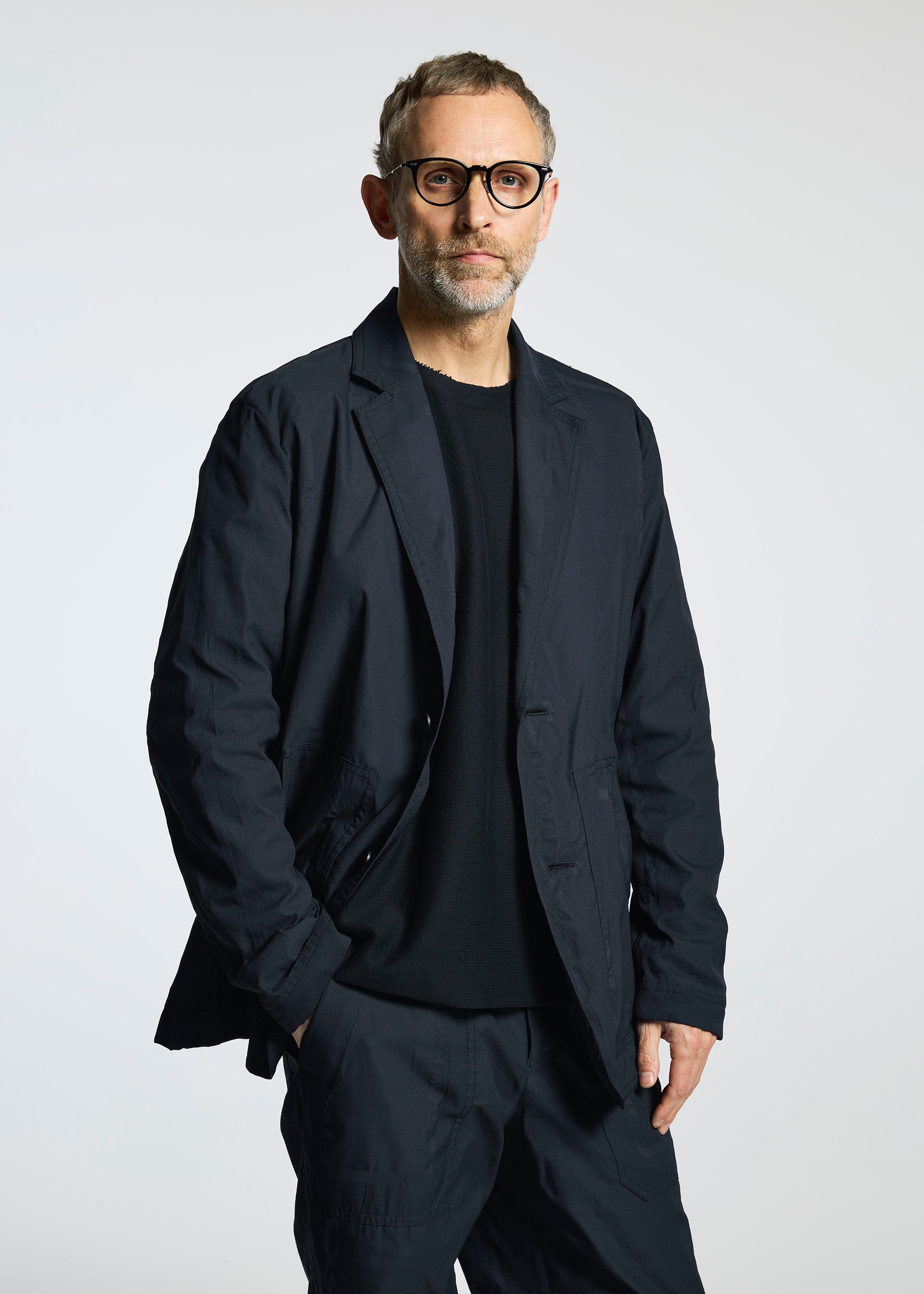 Zoomed view A man with short hair and a beard models the TYPE-U 001 from A-POC ABLE ISSEY MIYAKE—black glasses, wrinkle-resistant jacket, black shirt, and matching pants—standing before a plain light background, looking at the camera.