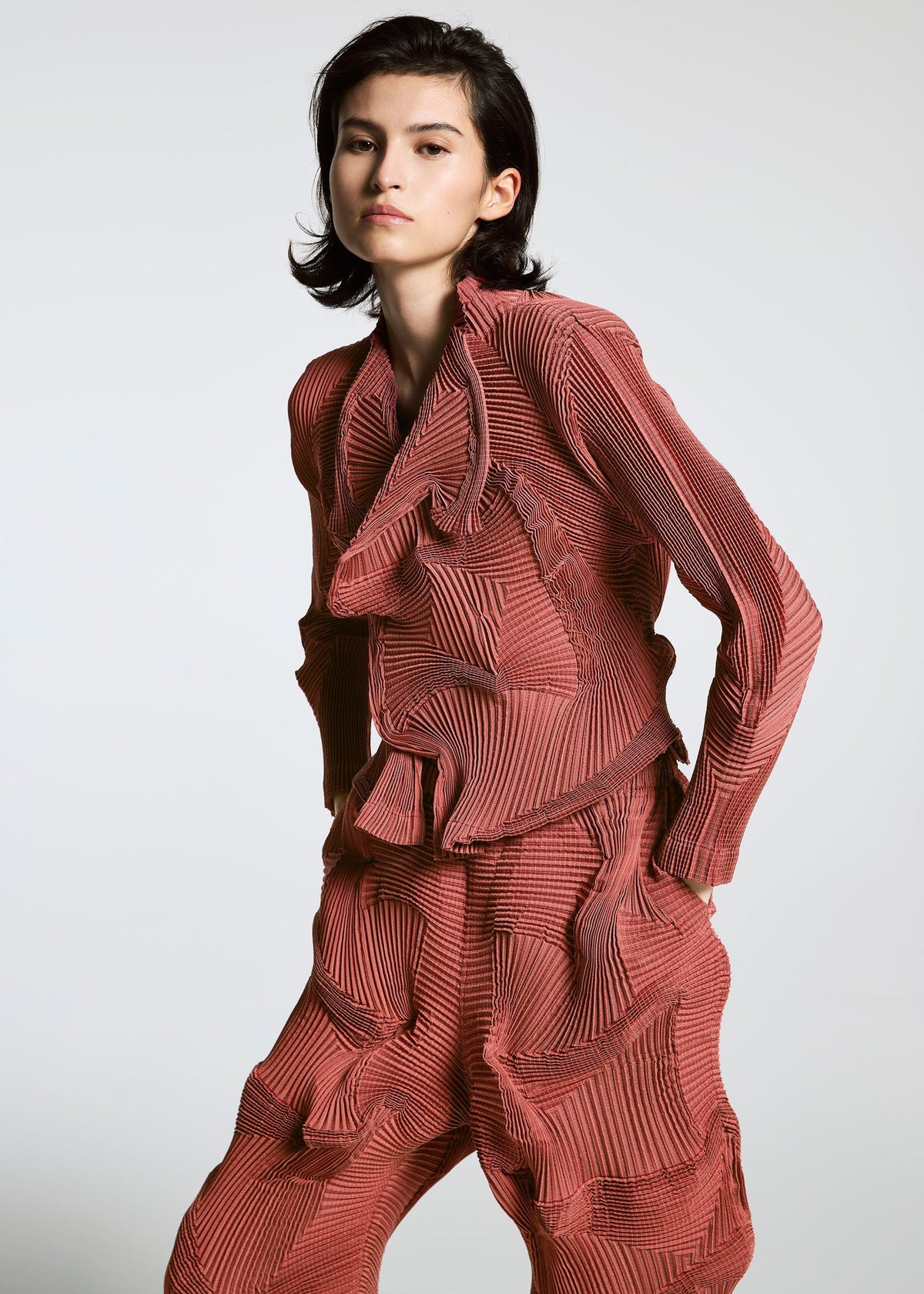 A person with short dark hair wears the TYPE-O 003 BK by A-POC ABLE ISSEY MIYAKE, a matching red textured set with a long-sleeve top and loose pants featuring wavy, sculpted details, standing against a plain light background. view 3 of 3