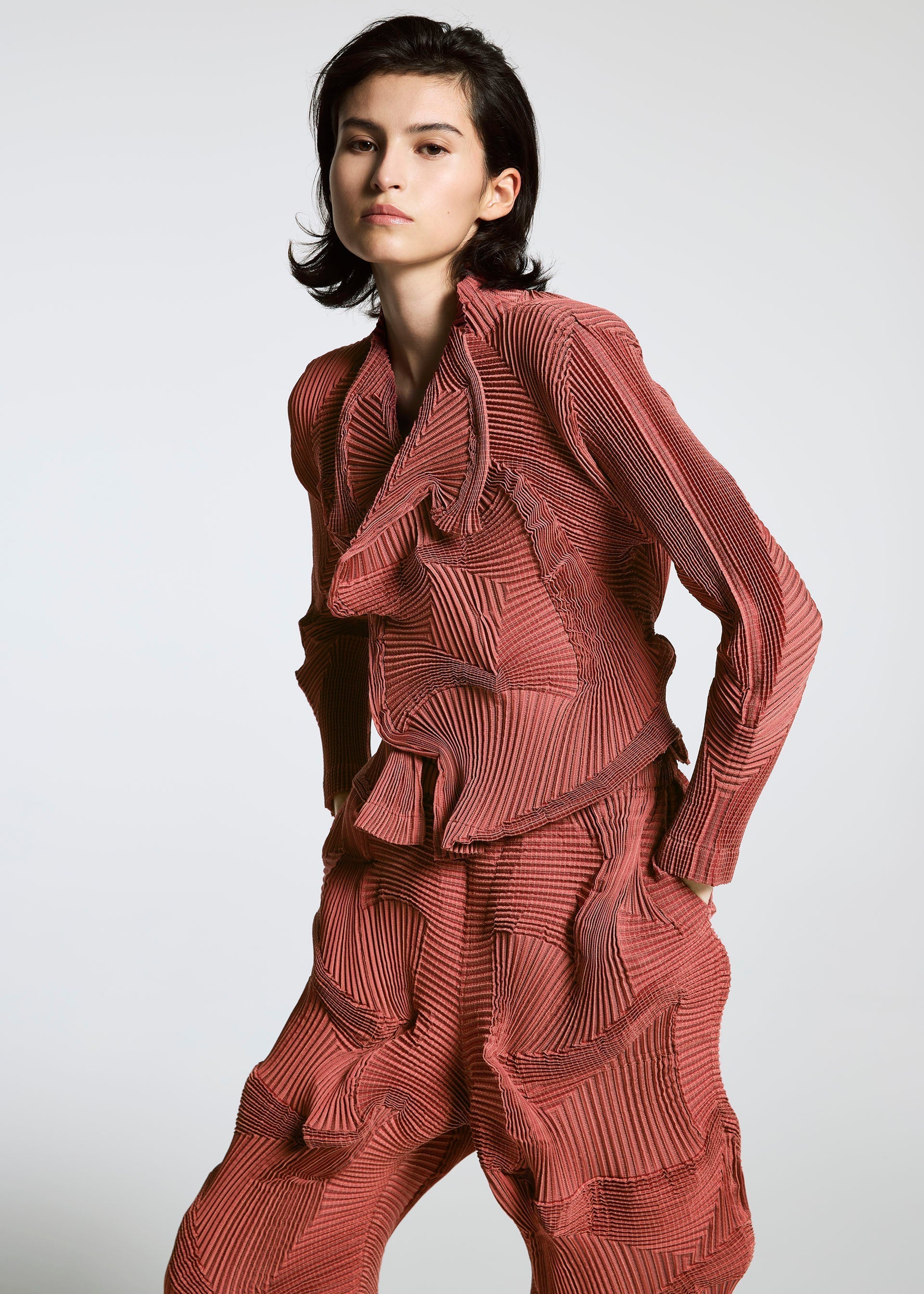 Zoomed view A person with short dark hair wears the TYPE-O 003 BK by A-POC ABLE ISSEY MIYAKE, a matching red textured set with a long-sleeve top and loose pants featuring wavy, sculpted details, standing against a plain light background.