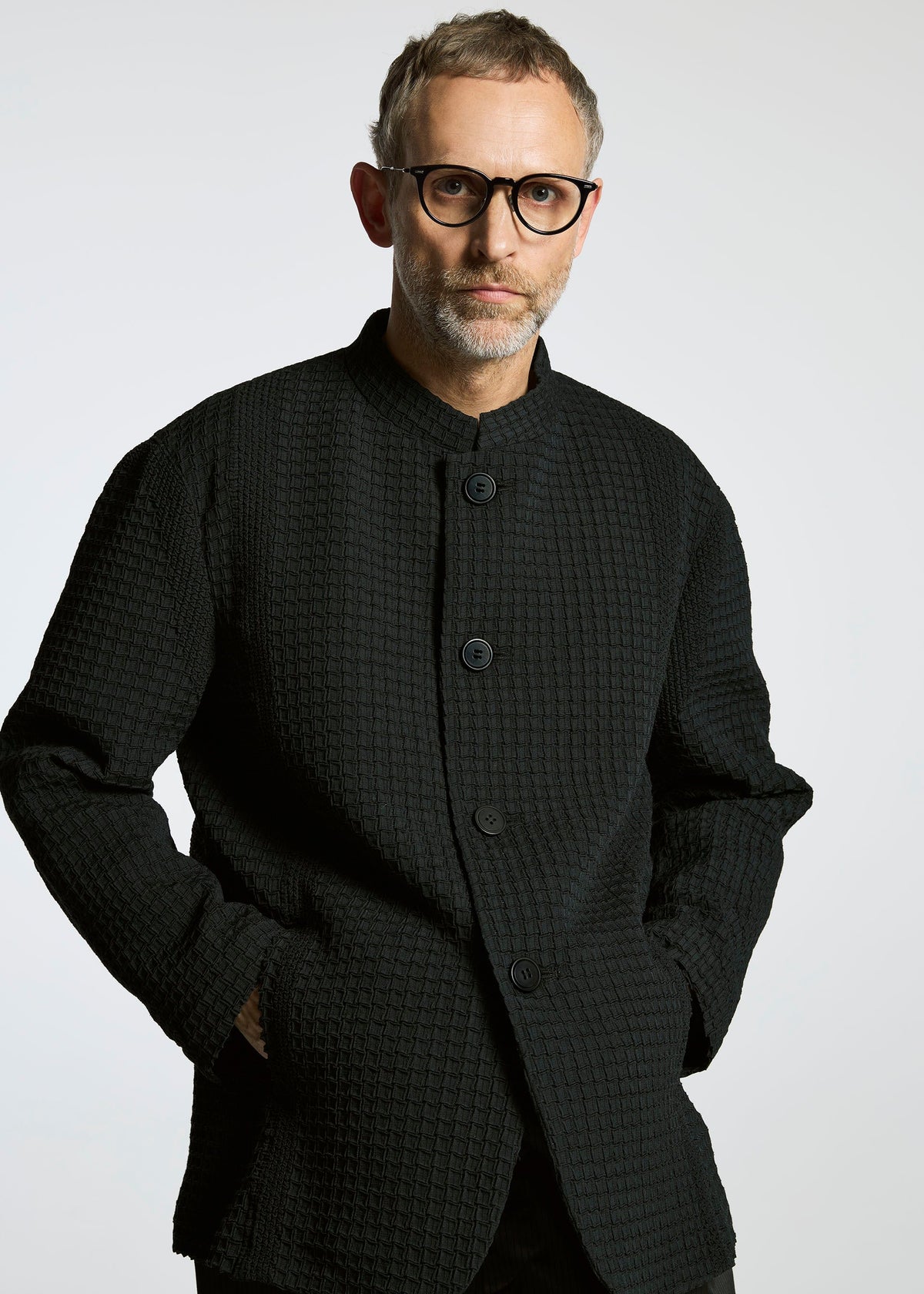 A man with short gray hair, a beard, and black-rimmed glasses wears the TYPE-O 014 standing collar jacket by A-POC ABLE ISSEY MIYAKE, posing with hands in pockets against a light gray background. view 5 of 6