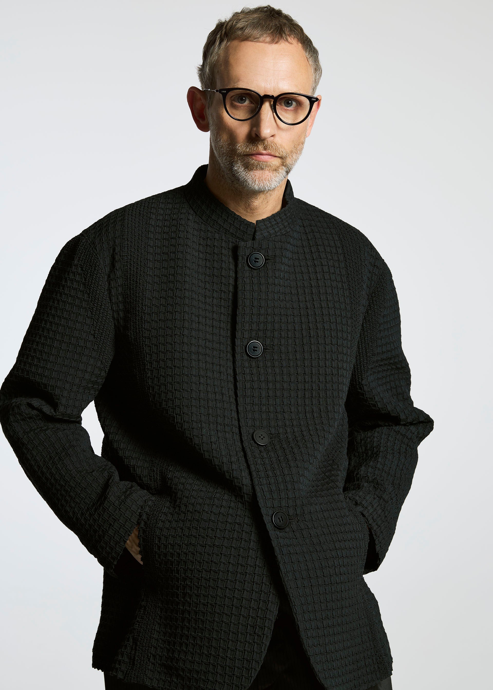 Zoomed view A man with short gray hair, a beard, and black-rimmed glasses wears the TYPE-O 014 standing collar jacket by A-POC ABLE ISSEY MIYAKE, posing with hands in pockets against a light gray background.