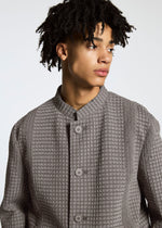 A young person with short, curly hair models the TYPE-O 014 standing collar jacket by A-POC ABLE ISSEY MIYAKE in textured light gray with subtle grid pleats, looking to the side against a plain background.
