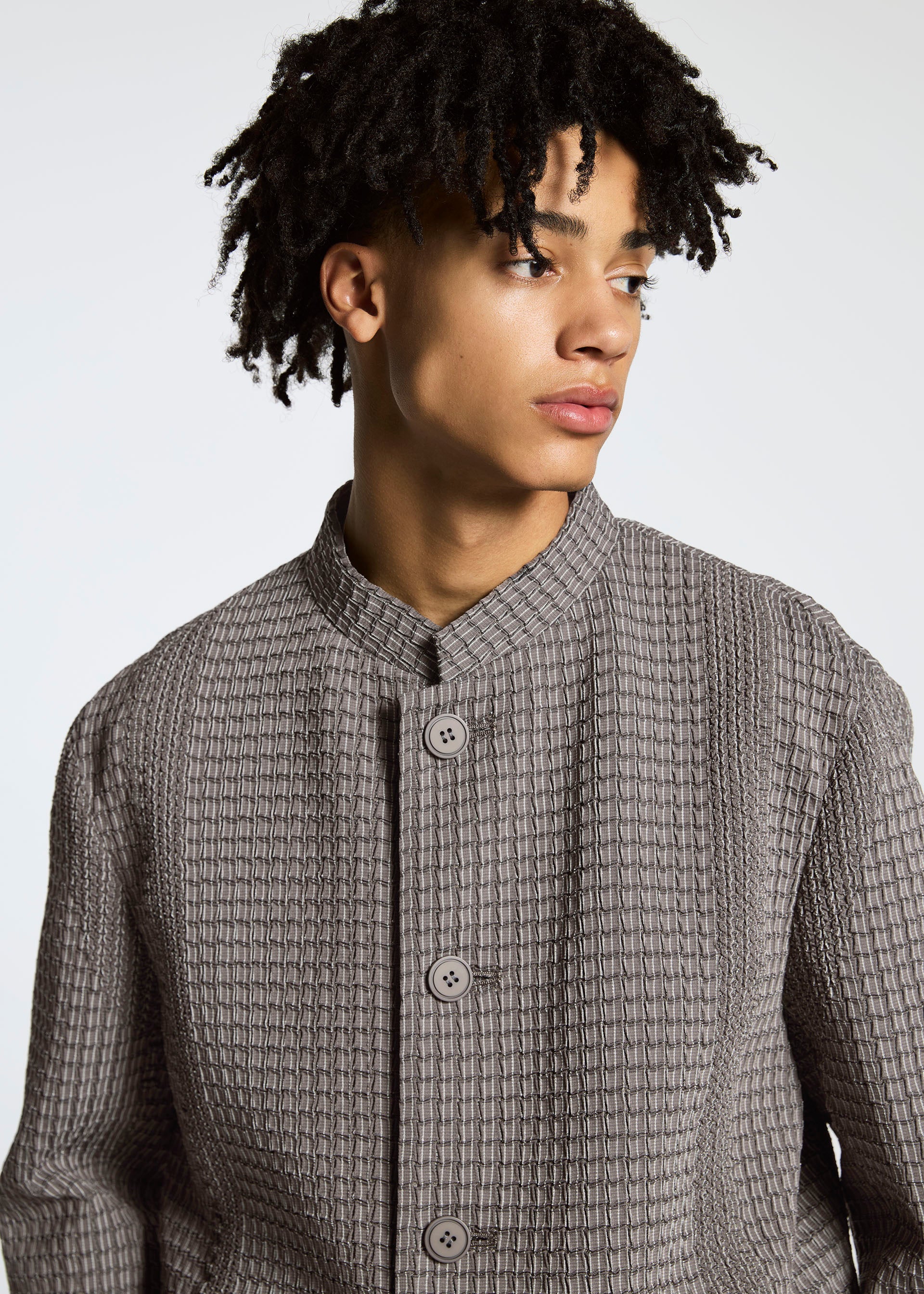Zoomed view A young person with short, curly hair models the TYPE-O 014 standing collar jacket by A-POC ABLE ISSEY MIYAKE in textured light gray with subtle grid pleats, looking to the side against a plain background.