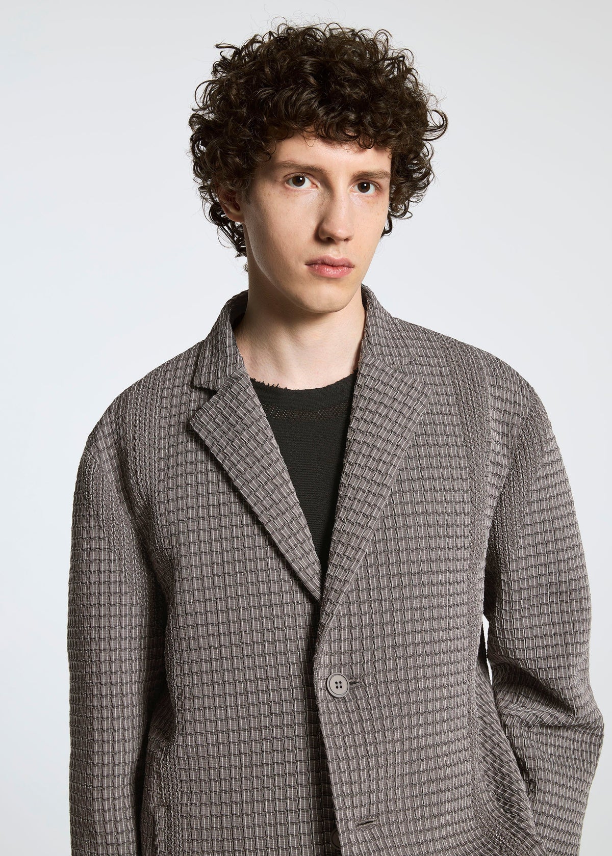 A young person with curly brown hair wears the TYPE-O 014 jacket by A-POC ABLE ISSEY MIYAKE, featuring grid-shaped pleats and a tailored Steam Stretch collar, standing against a plain light background and looking neutrally at the camera. view 5 of 5