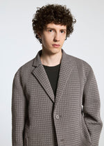 A young person with curly brown hair wears the TYPE-O 014 jacket by A-POC ABLE ISSEY MIYAKE, featuring grid-shaped pleats and a tailored Steam Stretch collar, standing against a plain light background and looking neutrally at the camera.