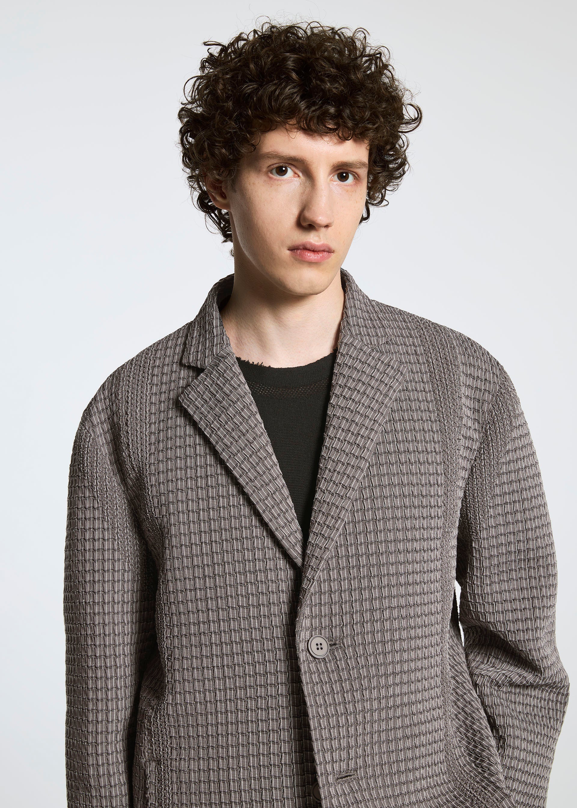 Zoomed view A young person with curly brown hair wears the TYPE-O 014 jacket by A-POC ABLE ISSEY MIYAKE, featuring grid-shaped pleats and a tailored Steam Stretch collar, standing against a plain light background and looking neutrally at the camera.