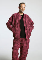 A man with short hair and a beard stands hands-in-pockets against a plain backdrop, wearing the TYPE-O 018 geometric maroon jacket and pleated pants by A-POC ABLE ISSEY MIYAKE over a black shirt.