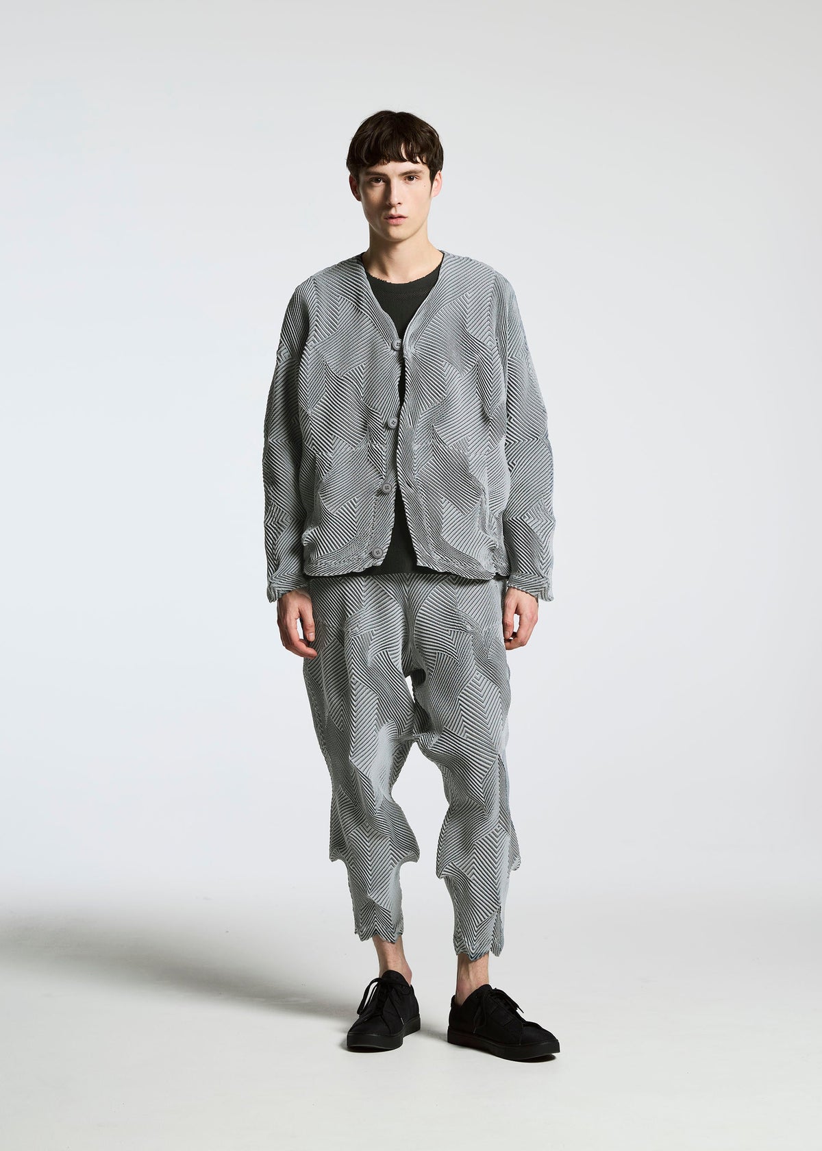A young man faces forward with a neutral expression, wearing A-POC ABLE ISSEY MIYAKE’s TYPE-O 010: a loose-fitting textured grey jacket and matching wavy pleated pants, paired with a black shirt and shoes, against a plain white background. view 5 of 11