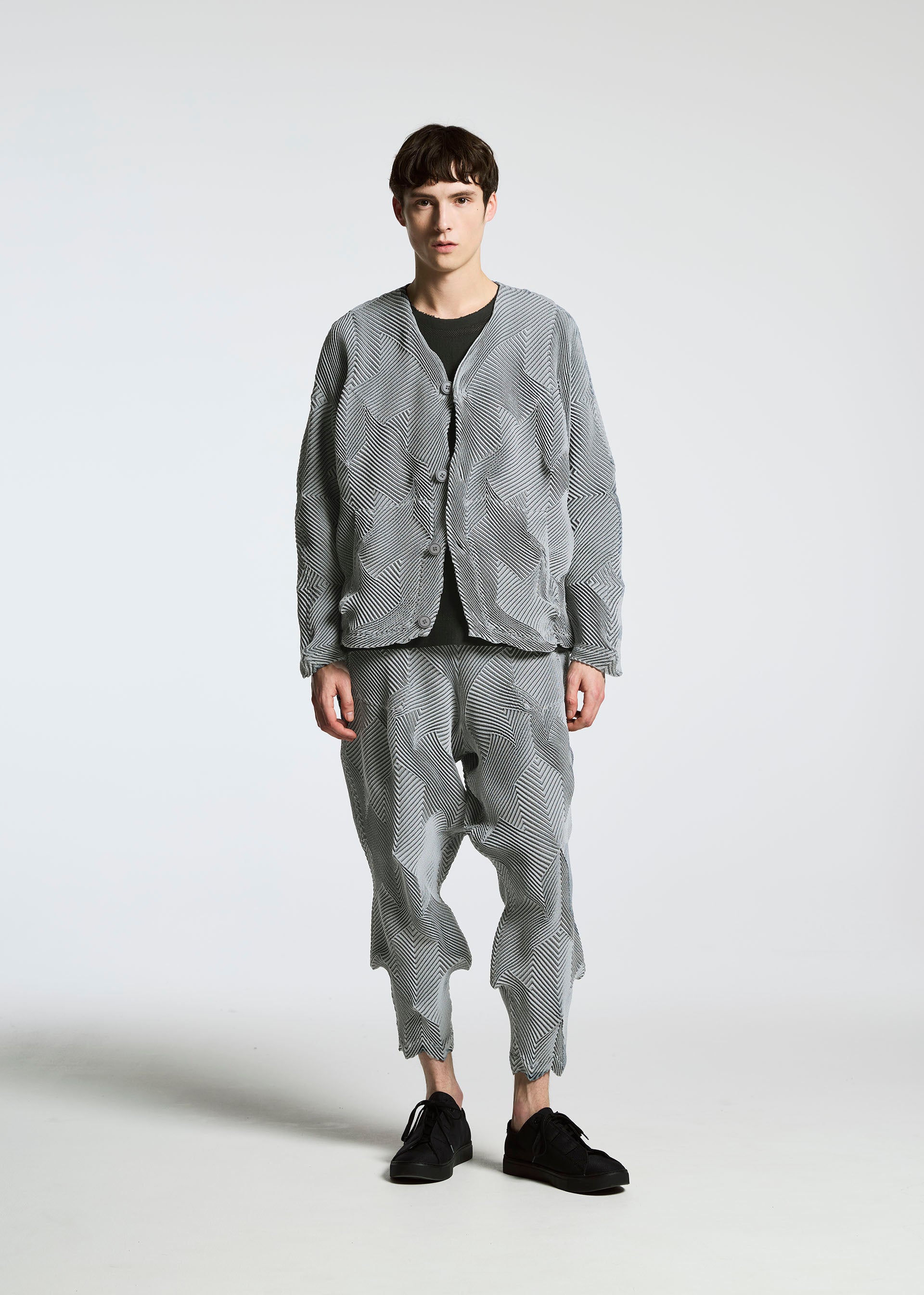 Zoomed view A young man faces forward with a neutral expression, wearing A-POC ABLE ISSEY MIYAKE’s TYPE-O 010: a loose-fitting textured grey jacket and matching wavy pleated pants, paired with a black shirt and shoes, against a plain white background.