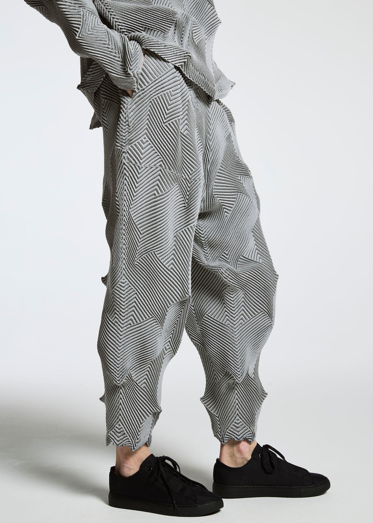 A person wears TYPE-O 010 loose, pleated gray pants with a star pattern and zigzag texture by A-POC ABLE ISSEY MIYAKE, made from recycled polyester, paired with black sneakers. The image is cropped at the shoulders to highlight the pants. view 6 of 11