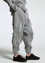 A person wears TYPE-O 010 loose, pleated gray pants with a star pattern and zigzag texture by A-POC ABLE ISSEY MIYAKE, made from recycled polyester, paired with black sneakers. The image is cropped at the shoulders to highlight the pants.
