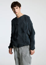 A young person with short brown hair wears the TYPE-O 010 by A-POC ABLE ISSEY MIYAKE— a loose, textured dark blue sweater with geometric patterns and light gray pleated houndstooth pants—against a plain white background.