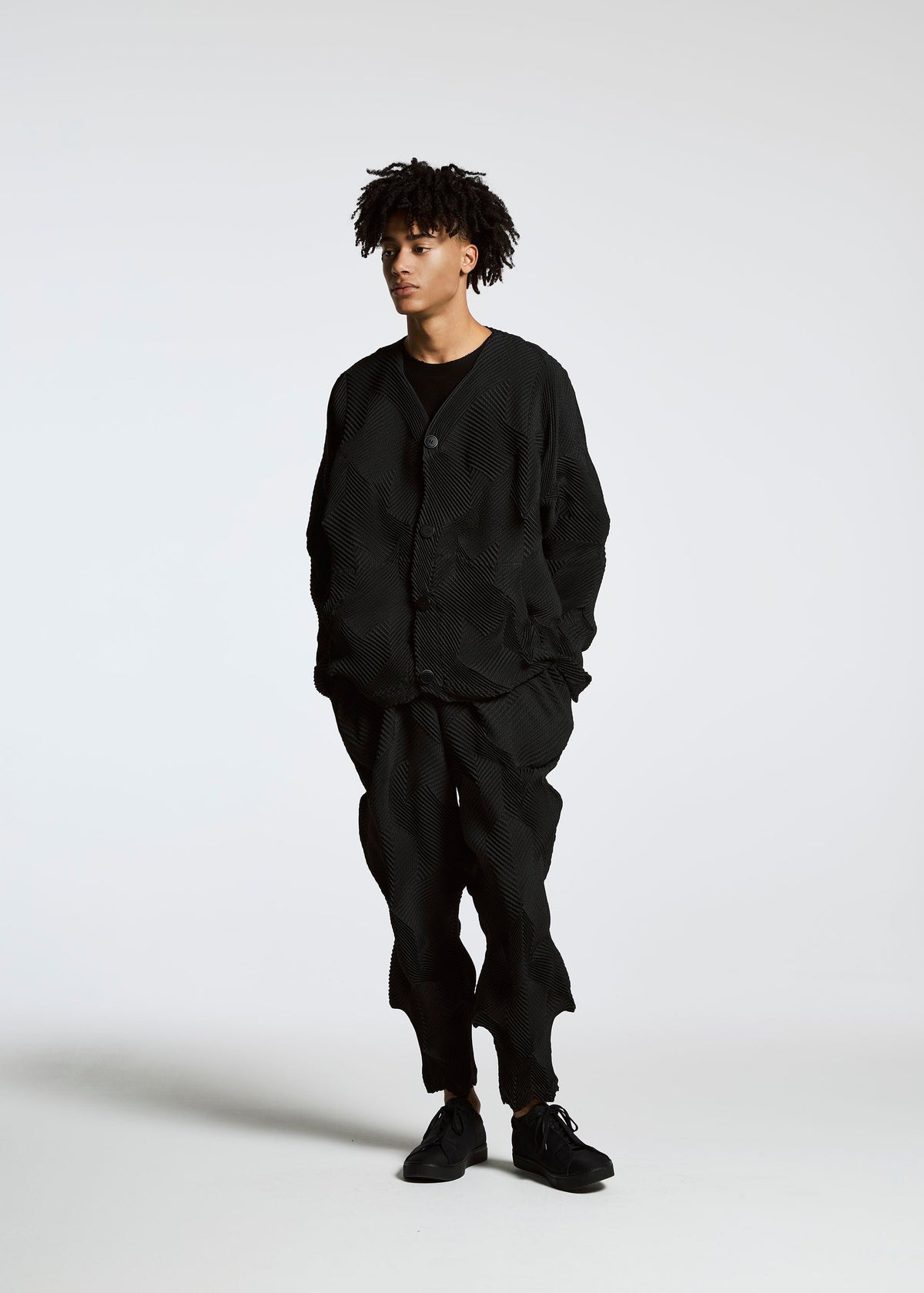 A person with short curly hair stands against a white background, wearing the TYPE-O 010 jacket and pants by A-POC ABLE ISSEY MIYAKE in black houndstooth, with hands in pockets and a relaxed posture. view 9 of 11