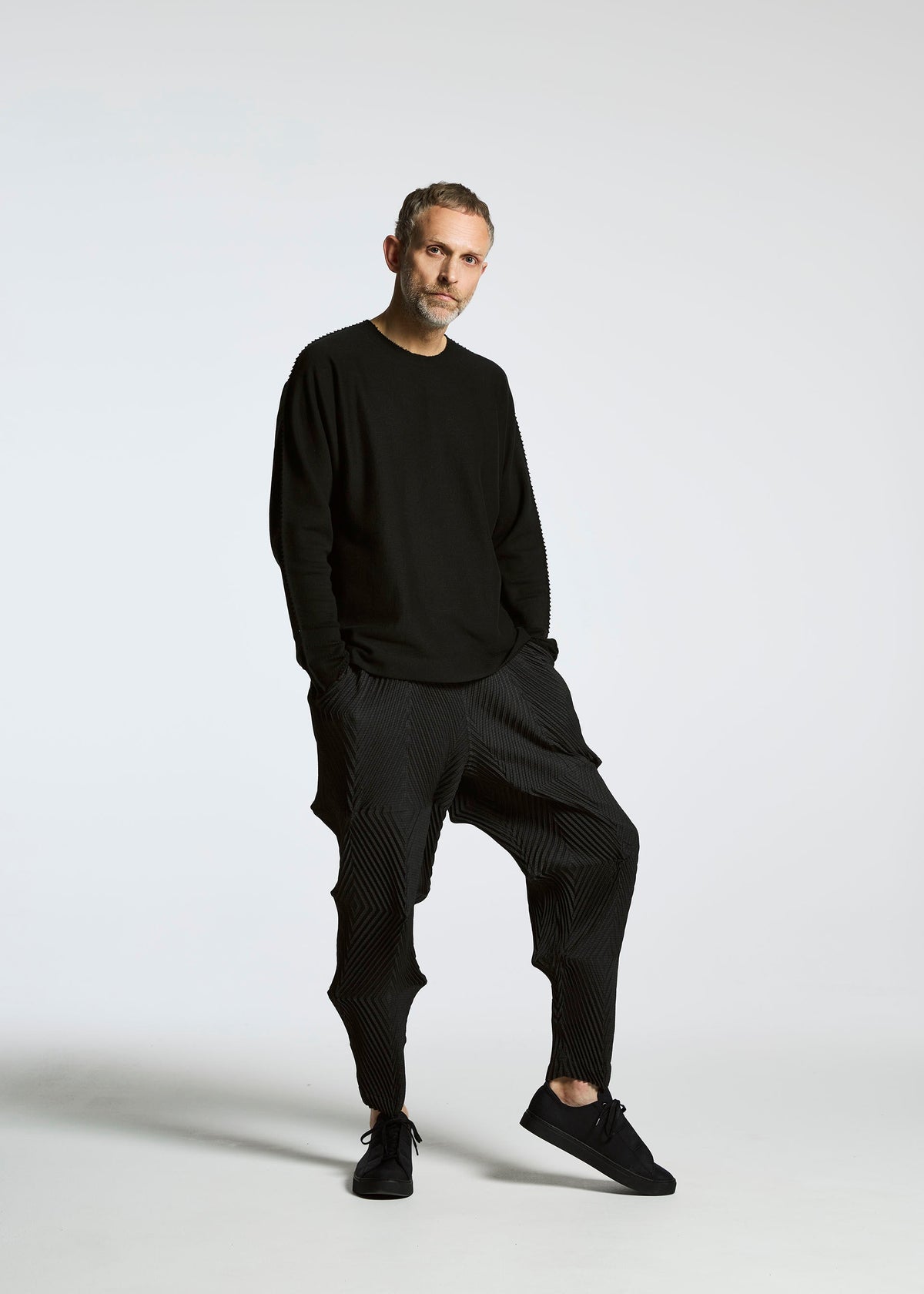 A man with short hair and a beard poses against a white background, wearing the TYPE-O 007 black long-sleeve shirt by A-POC ABLE ISSEY MIYAKE, pleated pants, and black sneakers, with one leg bent and hands in his pockets. view 5 of 10