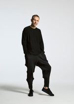 A man with short hair and a beard poses against a white background, wearing the TYPE-O 007 black long-sleeve shirt by A-POC ABLE ISSEY MIYAKE, pleated pants, and black sneakers, with one leg bent and hands in his pockets.