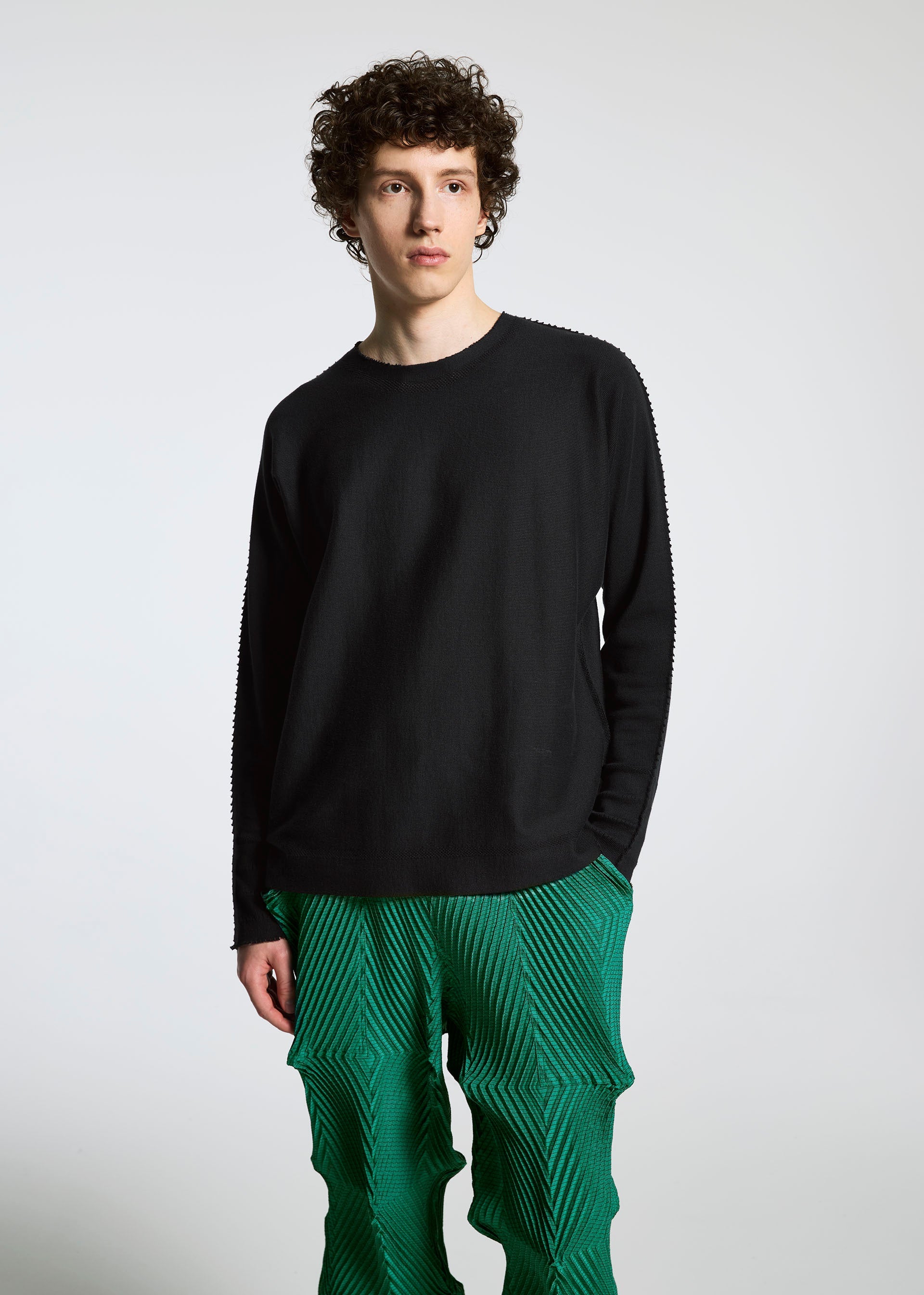Zoomed view A young person with curly hair poses against a plain background, dressed in a black long-sleeve shirt and vibrant green TYPE-O 007 Steam Stretch pants by A-POC ABLE ISSEY MIYAKE, featuring a striking geometric pattern.