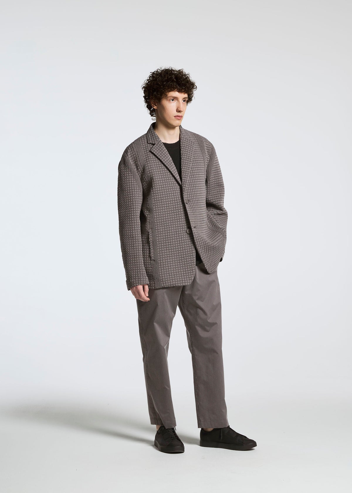 Wearing the TYPE-S 003 by A-POC ABLE ISSEY MIYAKE, a person with curly brown hair stands against a white background, dressed in a checked gray blazer and pants, black shirt and shoes, one hand in pocket and a neutral expression. view 5 of 15