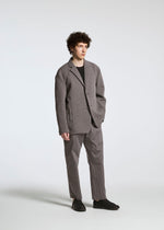 Wearing the TYPE-S 003 by A-POC ABLE ISSEY MIYAKE, a person with curly brown hair stands against a white background, dressed in a checked gray blazer and pants, black shirt and shoes, one hand in pocket and a neutral expression.