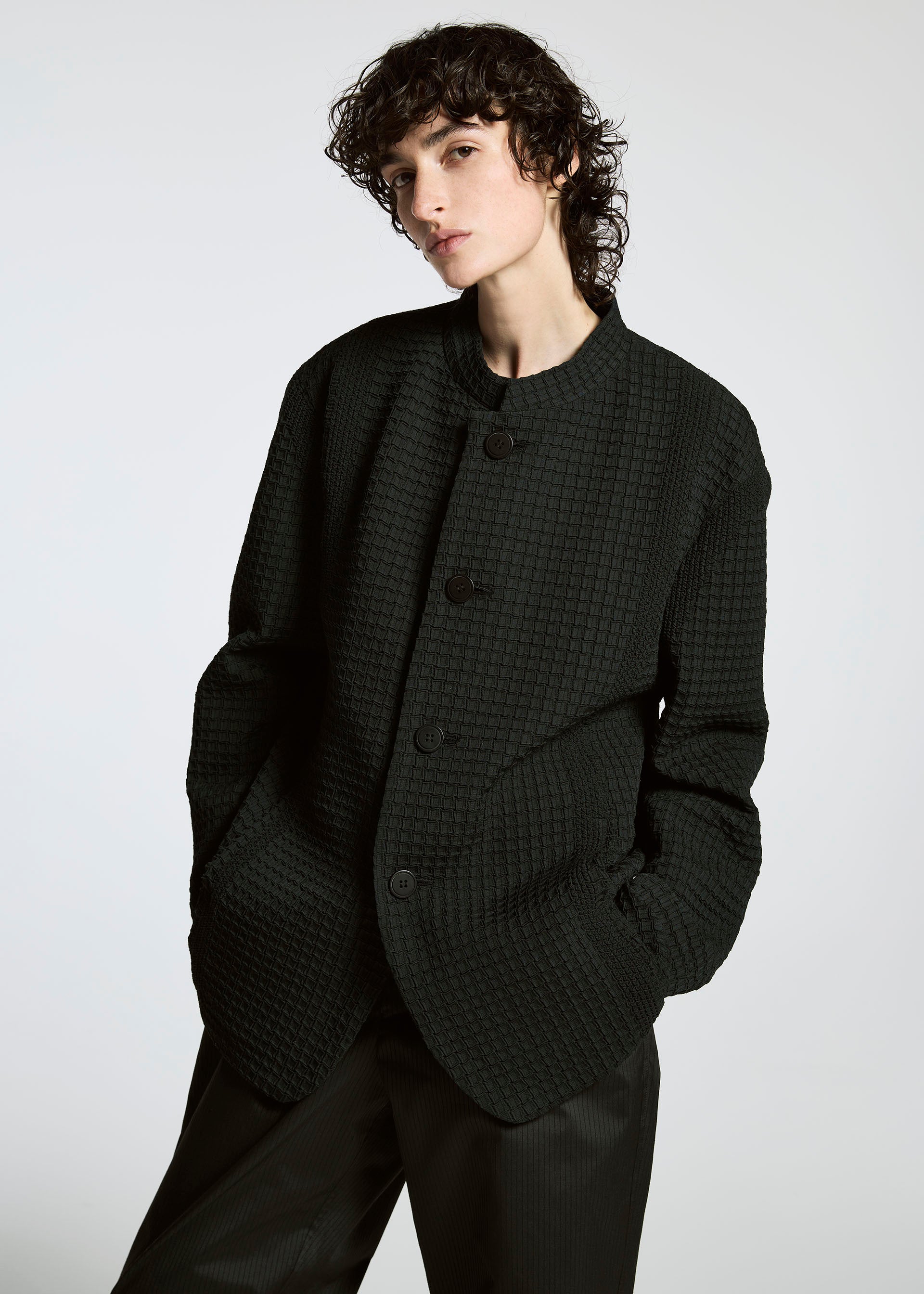 Zoomed view Wearing the TYPE-S 003 by A-POC ABLE ISSEY MIYAKE, a person with short, curly dark hair stands against a plain light background in a textured oversized black button-up jacket and lightweight stretchy pants, hands in pockets.