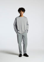 A person with short dreadlocks and a neutral expression wears the TYPE-C 003 by A-POC ABLE ISSEY MIYAKE: a loose, light gray pleated shirt and matching cotton pants, styled with black shoes, against a plain white background.