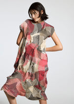 A person with short dark hair models the TYPE-P 002 dress by A-POC ABLE ISSEY MIYAKE, featuring loose pleats and geometric patterns in vivid red, black, and beige against a plain light background.