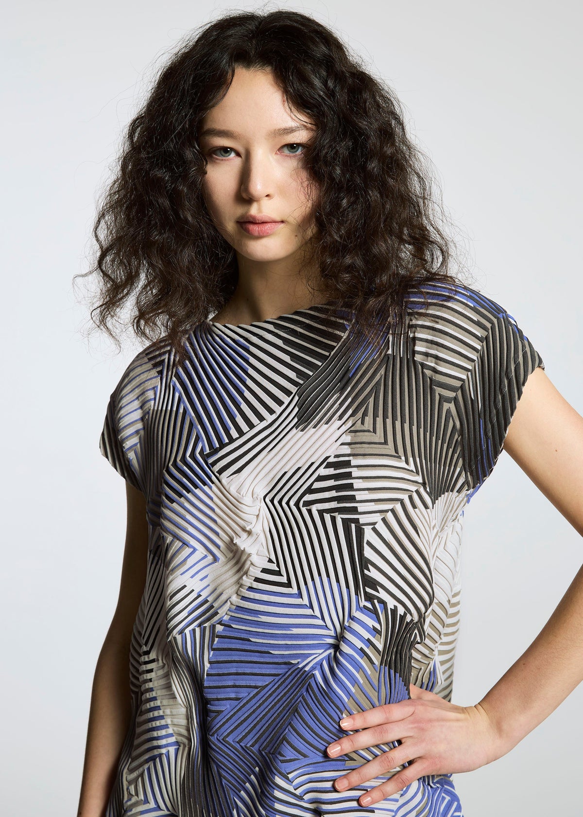 A woman with wavy dark hair poses against a plain light gray background, wearing the TYPE-P 002 short-sleeve geometric-patterned shirt in vivid black, white, and blue by A-POC ABLE ISSEY MIYAKE. view 5 of 6