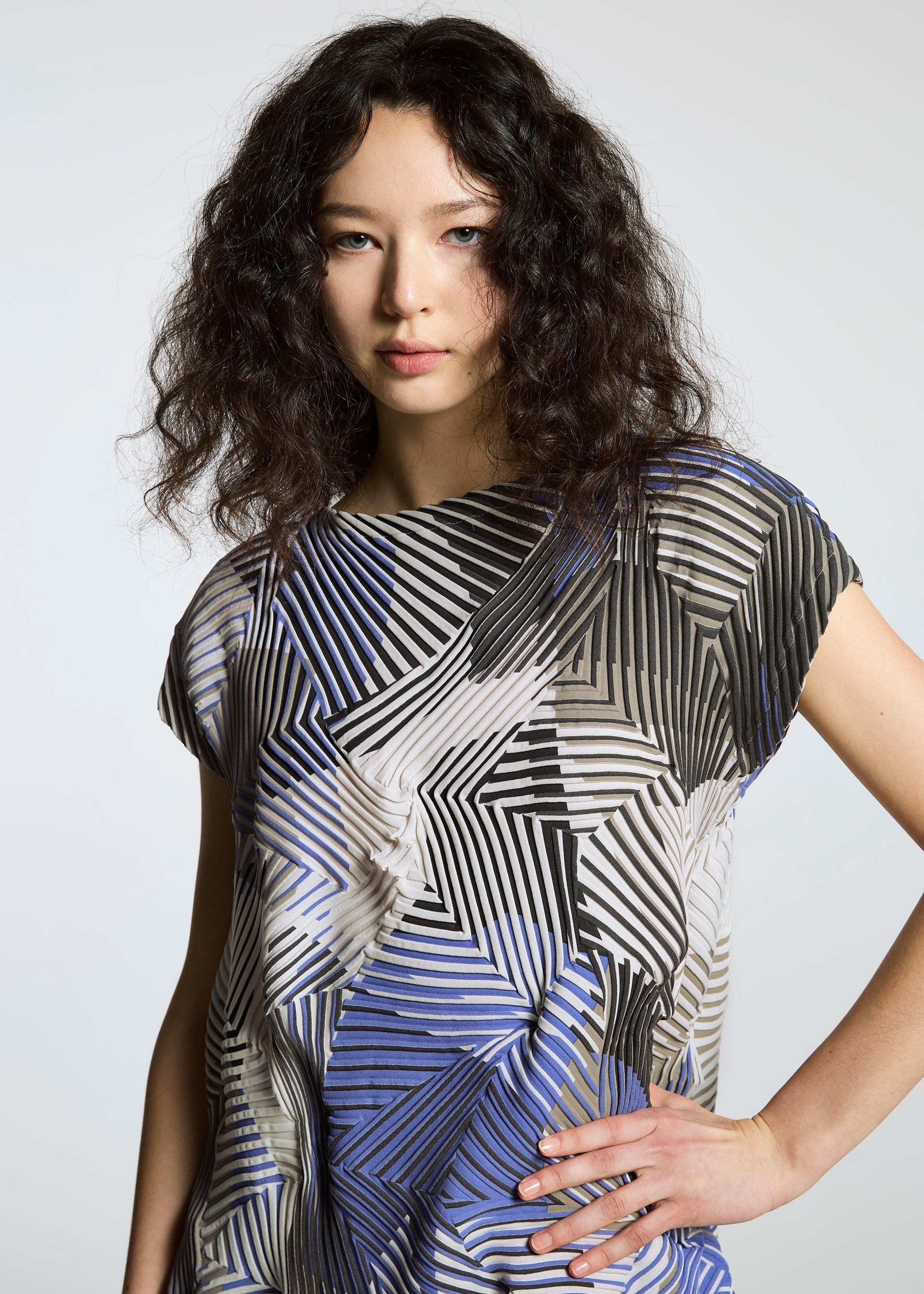 Zoomed view A woman with wavy dark hair poses against a plain light gray background, wearing the TYPE-P 002 short-sleeve geometric-patterned shirt in vivid black, white, and blue by A-POC ABLE ISSEY MIYAKE.