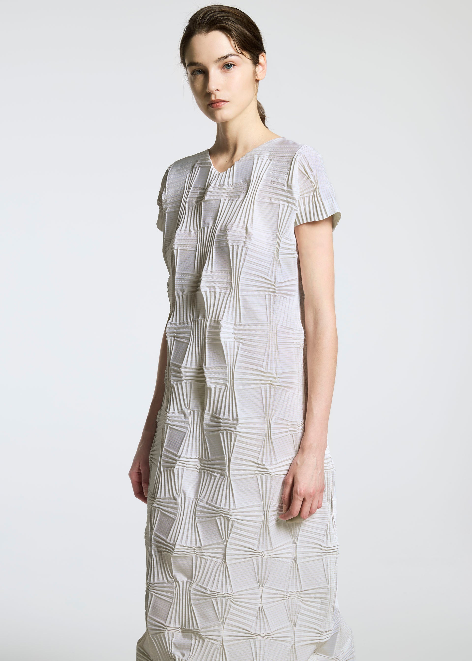 Zoomed view A woman models the TYPE-O 018 dress by A-POC ABLE ISSEY MIYAKE, featuring a textured geometric spiral pattern in white and beige, standing against a plain light gray background and gazing calmly at the camera.