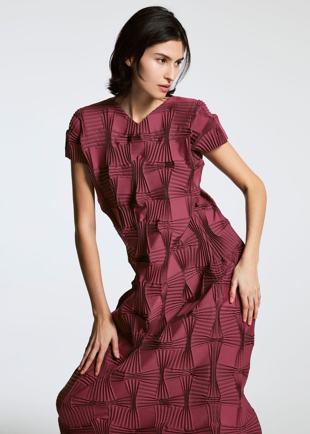 Someone poses against a plain background wearing the TYPE-O 018 short-sleeved, textured maroon dress with geometric raised square patterns and steam stretch detailing by A-POC ABLE ISSEY MIYAKE. view 5 of 6