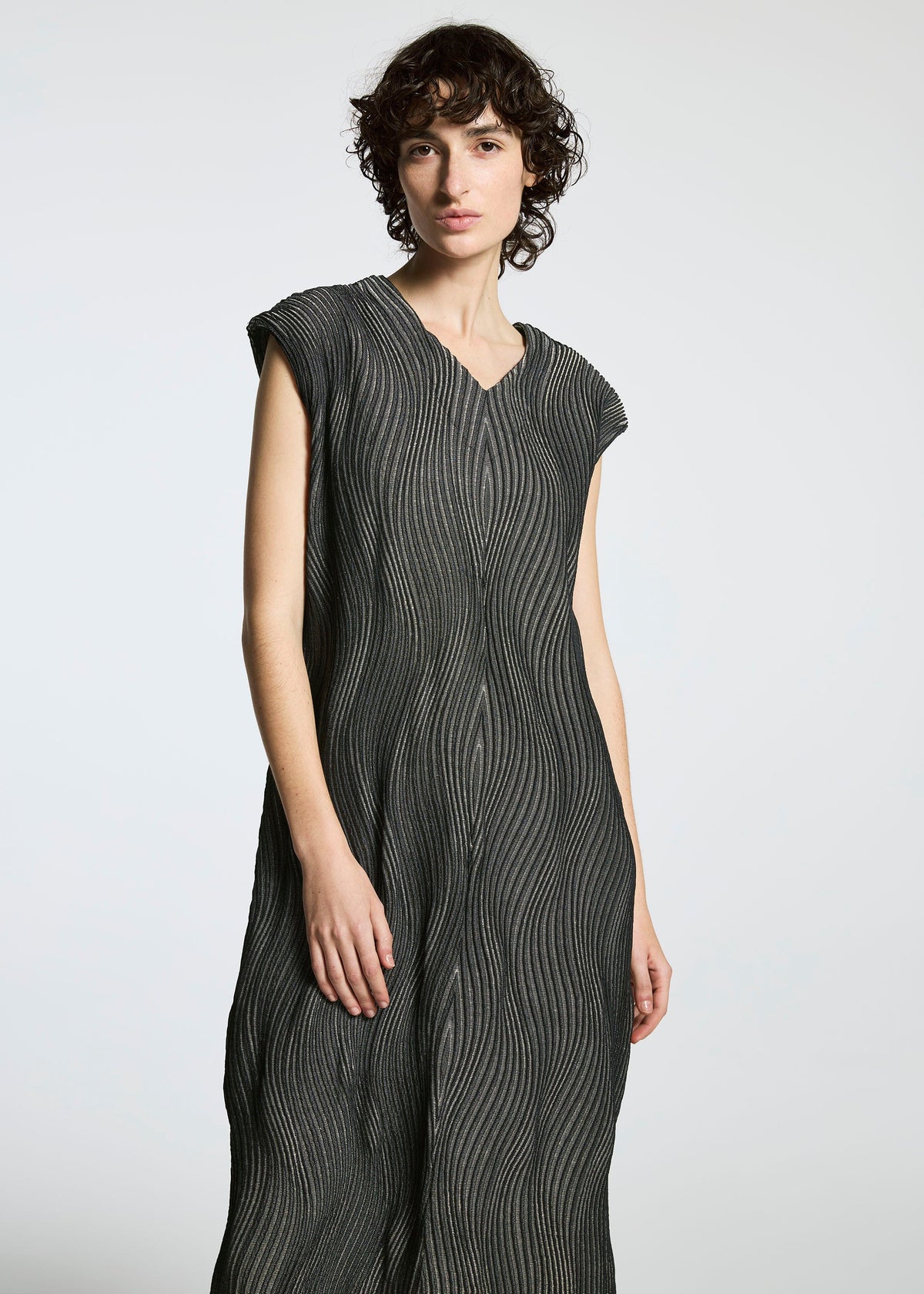 A person with short, curly hair wears a textured, sleeveless gray dress from the TYPE-XI Spiber project by A-POC ABLE ISSEY MIYAKE, made with Brewed Protein fiber and featuring subtle vertical wavy lines against a plain background. view 3 of 7