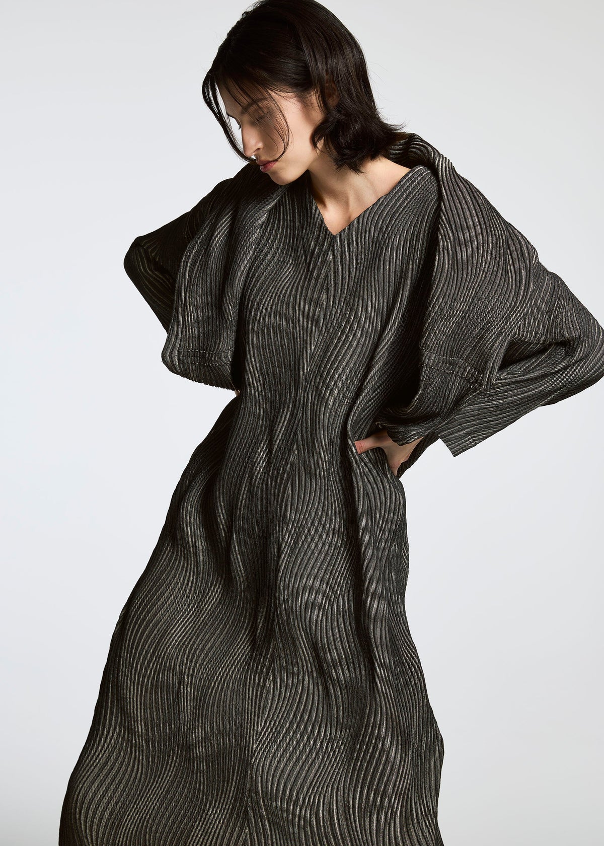 A person with short dark hair wears a long, textured, dark dress featuring wavy vertical lines, crafted from Brewed Protein fiber. The garment is the TYPE-XI Spiber project by A-POC ABLE ISSEY MIYAKE. They pose against a plain light background. view 4 of 7