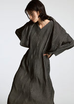 A person with short dark hair wears a long, textured, dark dress featuring wavy vertical lines, crafted from Brewed Protein fiber. The garment is the TYPE-XI Spiber project by A-POC ABLE ISSEY MIYAKE. They pose against a plain light background.