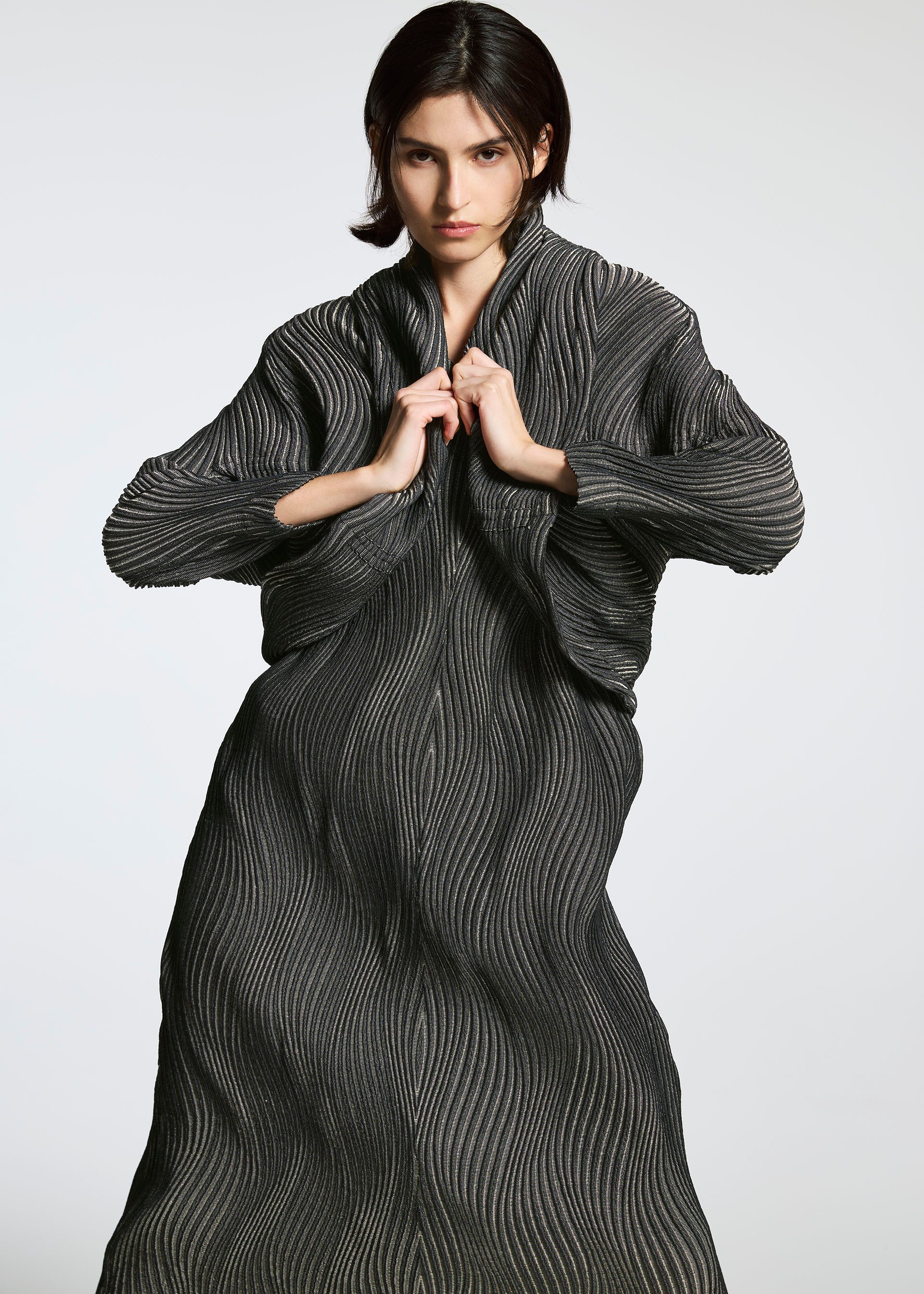Zoomed view A person with dark hair poses in a long, textured dark gray robe dress made from Brewed Protein fiber by A-POC ABLE ISSEY MIYAKE, TYPE-XI Spiber project, holding the collar with both hands against a plain light background.