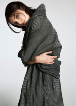 A person with dark hair wears a textured, wavy, gray and black pleated TYPE-XI Spiber project garment by A-POC ABLE ISSEY MIYAKE, posing with arms crossed and looking at the camera against a plain light background.